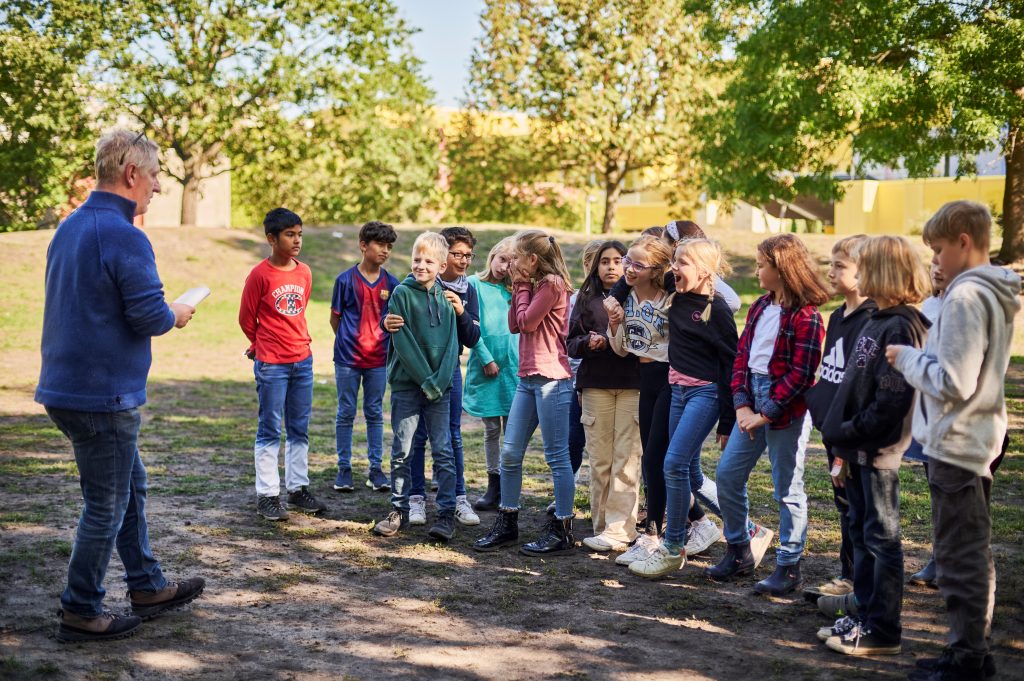 Eine Gruppe von Kindern mit einer Lehrperson steht auf einer Wiese.